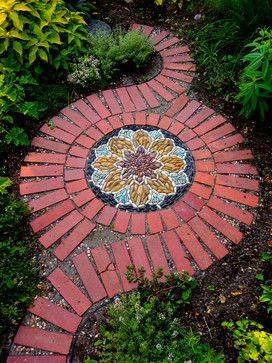 Gartenbau Gartengestaltung Landschaftsgestaltung Pflasterarbeiten Ein geschwungener Weg aus roten Ziegeln mit einem bunten Mosaik in der Mitte, umgeben von Pflanzen.