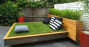 Gartenbau Gartengestaltung Landschaftsgestaltung Sitzfläche mit Gras und Kissen, umgeben von Holz und Pflanzen in einem Garten.